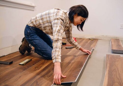 A woman is on her knees, laying down a piece of laminate flooring during installation